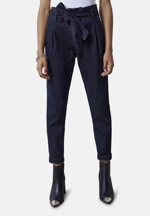 Kobieta ubrana w ciemnoniebieskie spodnie o wysokim stanie i zwężanych nogawkach, zawiązane paskiem z tkaniny, z mankietami na dole oraz czarne botki z metalowymi noskami.