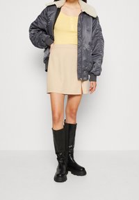 Chaqueta bomber gris con cuello de borrego, camiseta sin mangas amarilla de canalé, mini falda beige y botas altas de cuero negras.