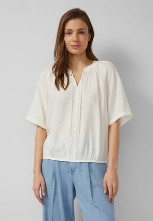 Jeune femme aux longs cheveux bruns portant un chemisier blanc ample à manches courtes et un jean taille haute bleu clair sur un fond uni.