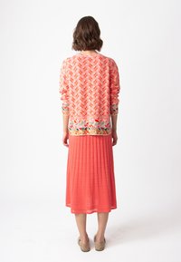 Korallmönstrad cardigan med långa ärmar och en ensfärgad korallfärgad plisserad maxikjol. Texturerad tyg, blommig dekoration vid cardiganens nederkant.