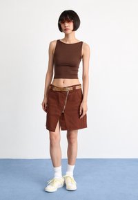 Braunes, figurbetontes Tanktop, hoch taillierter brauner Rock mit vorderem Reißverschluss und Schlitz, genietetem beige-gelbem Gürtel, kombiniert mit gelben Sneakers und weißen Socken.