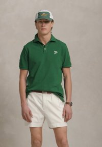 Cămașă polo verde cu un logo mic, pantaloni scurți albi și o șapcă bicromatică cu un logo. Accesorii includ o brățară și o centură.