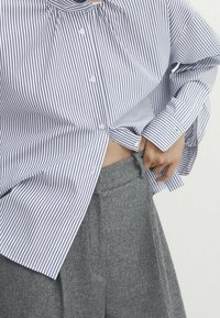 Chemise boutonnée à rayures bleues et blanches, avec un col froncé et des manches longues, associée à un pantalon taille haute gris.