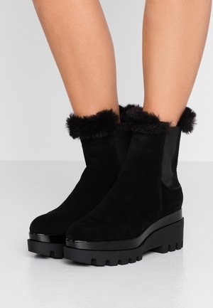 Bottines noires à semelles épaisses avec plateformes et bordures en fourrure portées sur des jambes nues devant un fond uni.