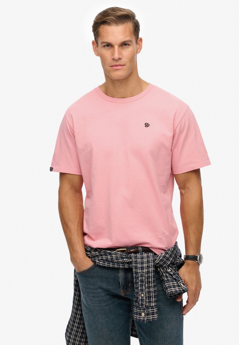 Roze katoenen t-shirt met korte mouwen, met een klein zwart logo op de borst. Gekörd met donkere jeans en een geruite blouse om de taille geknoopt.