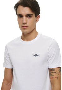 T-shirt bianco in cotone con scollo rotondo, maniche corte e logo di aquila ricamato in blu navy sul petto. Texture liscia, vestibilità rilassata.