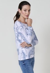 Top blanco de manga larga con un estampado de paisley azul, cuello redondo y un corte relajado; combinado con jeans azul claro.