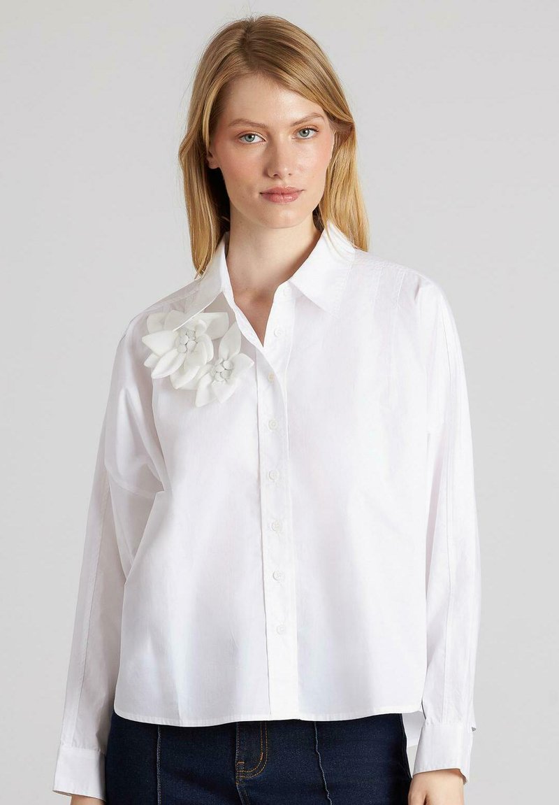 Camisa blanca de algodón con botones, que presenta un acento floral, corte relajado, mangas largas y una textura suave. Botones que recorren la parte delantera.