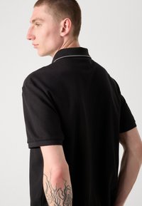 Svart pikétröja med korta ärmar och en kontrasterande grå krage. Har en texturerad tygyta och en figurnära design.