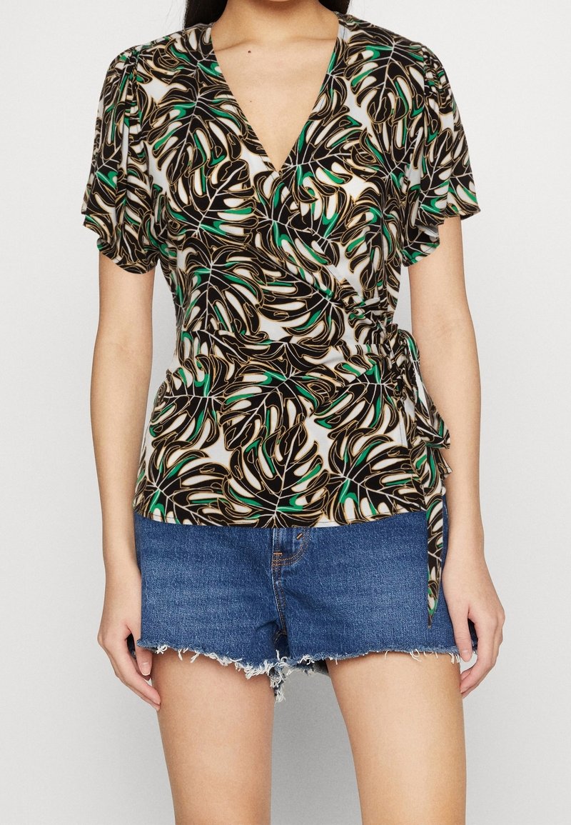 Femme portant un chemisier manches courtes, style portefeuille, avec un motif de feuilles noir, blanc, vert et or, associé à un short en denim effiloché.
