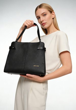 Femme blonde en tenue crème tenant un sac à main en cuir noir Pierre Cardin avec double anses sur fond blanc.