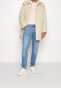 Man som bär en beige jacka över en beige och vit randig skjorta, ljusblå jeans och vita sneakers, står mot en vit bakgrund.