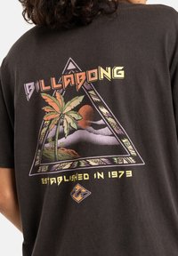Musta puuvillapaita, jossa on kolmiomainen graafinen muotoilu, jossa näkyy auringonlasku, palmun kuva ja "BILLABONG"-teksti värikkäällä, rohkealla fontilla.