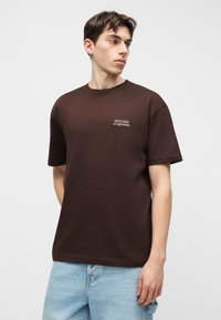 Joven con camiseta marrón con el texto "the new originals" mira hacia un lado, vistiendo vaqueros azul claro contra un fondo gris liso.