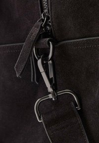 Detalle de un bolso de ante negro que muestra un clip metálico, un zipper suave y un sistema de sujeción texturizado. Presenta un hardware elegante y moderno.