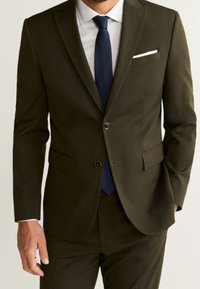 Abito verde oliva scuro con una vestibilità slim, giacca monopetto, rever a punta e due tasche. Abbinato a una camicia bianca e a una cravatta blu navy.