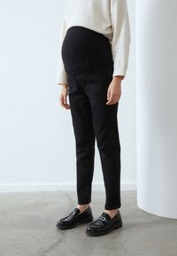 Mustad rasedusjeansid veniva vöötmega, kitseneva lõikega ja peene tekstuuriga, kombineeritud mustade loafide ja kreemja kampsuniga.