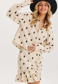 Béžový ľanový outfit s čiernym vzorom listov; obsahuje dlhý rukáv košele na gombíky a sukňu s volánovým ozdobením na leme.