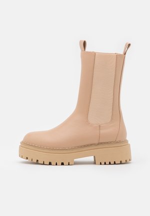 Beżowy skórzany botek za kostkę z elastycznymi panelami po bokach, zaokrąglonym noskiem oraz grubą beżową gumową podeszwą z teksturowanym bieżnikiem zapewniającym przyczepność.