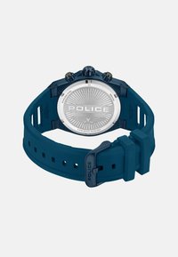 Blå gummirem klocka med klockkropp i rostfritt stål, med en texturerad silverfärgad urtavla, tre chronografsubskivor och "POLICE" graverat på baksidan.