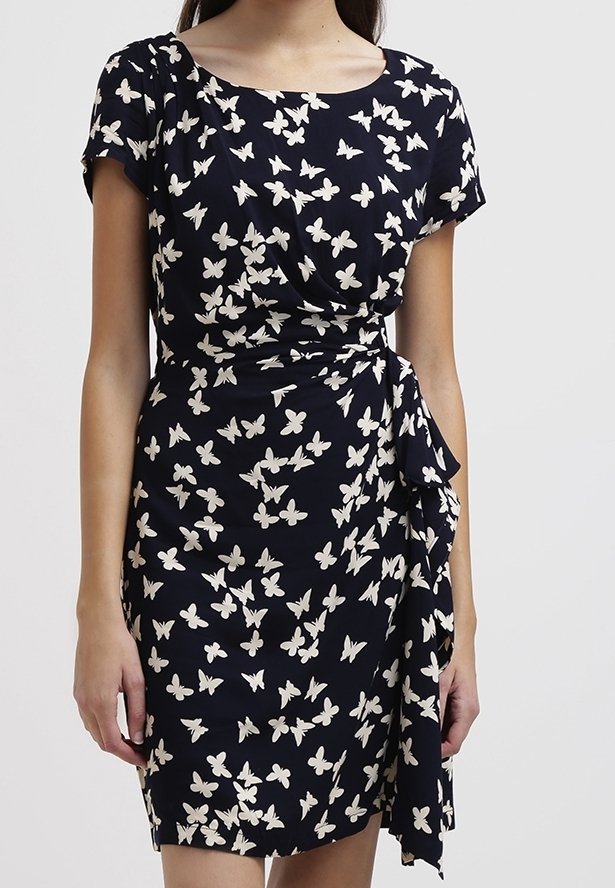 Femme portant une robe bleu marine à manches courtes avec un imprimé papillon blanc, dotée d’une taille froncée et d’un détail drapé sur le côté.