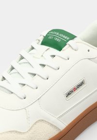 Weiße Sneaker mit einer strukturierten Lederoberfläche, Mesh-Akzenten, einem grünen Logo-Tag und brauner Gummisohle mit klassischer Schnürung.