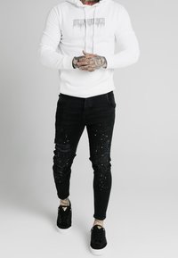 Vit huvtröja med ett grafiskt tryck, tillsammans med svarta skinny jeans med slitningar och splatterdetaljer. Svarta sneakers med vita sulor.