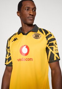 Κίτρινη αθλητική φανέλα με μαύρες λεπτομέρειες, που διαθέτει λογότυπο, κοντά μανίκια και κανονικό γιακά. Κατασκευασμένη από ελαφρύ συνθετικό υλικό.