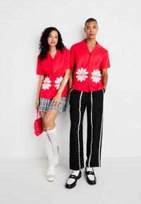 Červené tričká s krátkym rukávom s bielymi kvetinovými vzormi, skombinované s károvanými šortkami a čiernymi nohavicami s bielymi vlnkovanými akcentmi. Bielé ponožky a topánky.