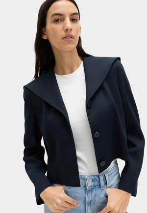 Jeune femme aux cheveux longs et foncés portant une veste courte bleu marine avec large col par-dessus une chemise blanche et un jean taille haute bleu clair.