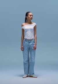 Baltas viršus be petnešėlių su sugrupuotomis detalėmis, derinamas su šviesiai mėlynais, plačiomis kelnėmis. Modelis dėvi baltus sportbačius, stovi prieš mėlyną foną.