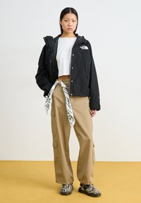 Musta vedenpitävä takki hupulla, vetoketjun edessä ja logolla. Valkoinen crop-top, beige cargo-housut taskuilla, ja mustat seepra-kuosiset kengät.