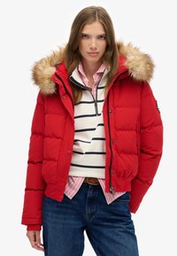 Doudoune rouge avec une capuche ornée de fourrure, fermeture éclair sur le devant et poignets élastiques, portée par-dessus une chemise rayée et un jean en denim bleu.