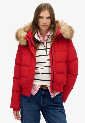 Red puffer jacket with a fur-trimmed hood, zippered front, and elastic cuffs, layered over a striped shirt and blue denim jeans.