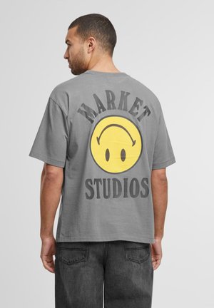 Grå bomullst-skjorte med et stort gult smilefjesgrafikk og "MARKET STUDIOS" tekst i fet svart på baksiden. Korte ermer, avslappet passform.