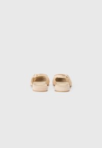 Beige szandálok, slip-on stílusúak, textúrázott fonott mintával, lapos talppal és sima felülettel. A hátsó nézet kerek formát és minimális részletezést mutat.