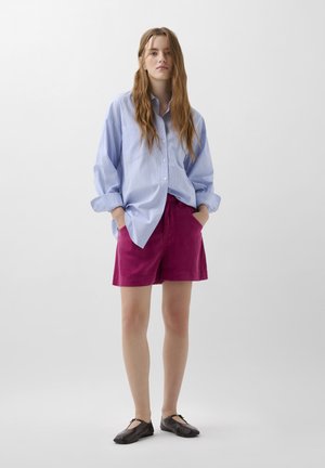 Modrá a bílá pruhovaná overshirt, růžové šortky a černé nazouvací boty. Tkanina vypadá hladce; šortky mají přední kapsy a volný střih.