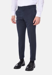 Pantaloni di ordinanza blu navy con frontale piatto, gambe affusolate e risvolti, caratterizzati da una texture liscia e passanti per cintura, abbinati a scarpe nere.