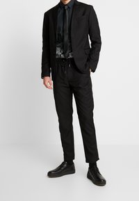 Black suit jacket over a black patterned shirt, paired with black drawstring trousers and black shoes. Sleek, tailored design.