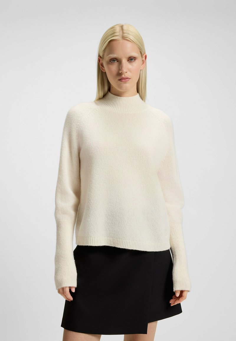 Maglione lavorato a maglia color crema con un alto collo a scatola e polsini a costine, abbinato a una gonna nera. Texture morbida e vestibilità rilassata.