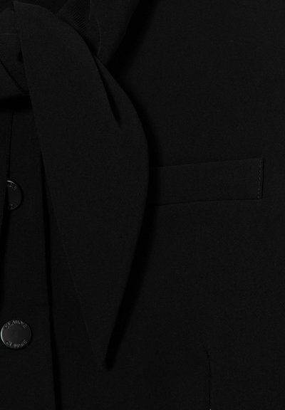Blazer nero con dettaglio della cravatta in tessuto coordinato e bottoni visibili sul davanti a sinistra.