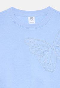 Gaiši zils džemperis ar tauriņa dizainu izšūts kontrastējošā diegā, ar apļveida kakla izgriezumu un mīkstu tekstūru. Redzams GAP Kids uzraksts.