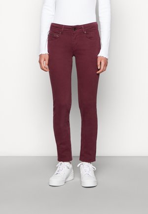 Woman wearing slim-fit dark red jeans, white long-sleeve top, and white platform sneakers, standing against a plain light background.