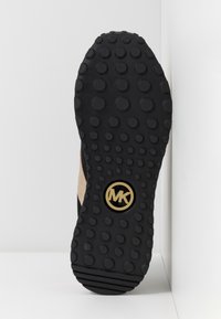 Semelle de chaussure noire avec des motifs circulaires et un logo MK doré au centre, affichée sur fond blanc.