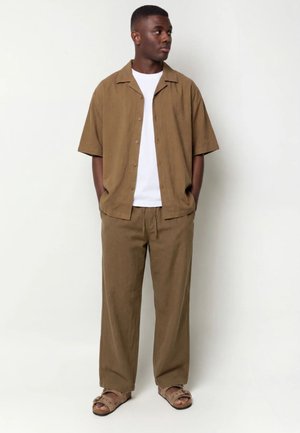 Mann trägt braunes kurzärmeliges Hemd mit Knöpfen, passende braune Hose, weißes T-Shirt und braune Sandalen, steht mit Händen in den Taschen und schaut nach rechts.