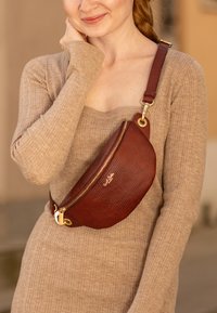 Borsa a tracolla in pelle marrone con finitura texturizzata, chiusura a zip e dettagli in oro, indossata su un vestito lungo a maniche lunghe beige a coste.