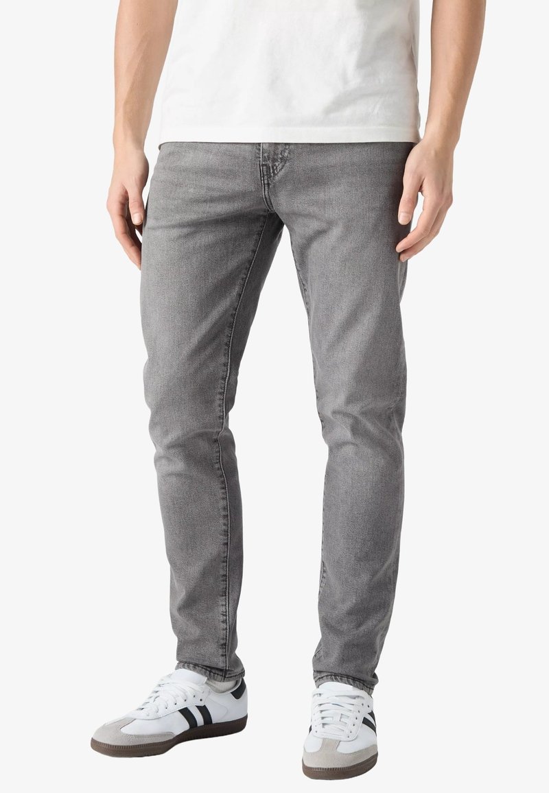 Homme portant un jean gris slim, des baskets blanches à bandes noires et un t-shirt blanc, debout devant un fond uni.
