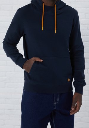 Mann trägt dunkelblauen Hoodie mit orangenen Kordeln, Hand in der vorderen Tasche, kombiniert mit dunkelblauen Jeans vor einer weißen Ziegelwand.