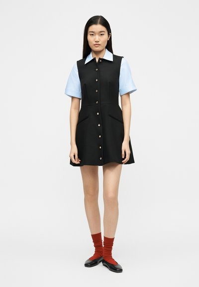 Γυναίκα που στέκεται φορώντας μαύρο αμάνικο φόρεμα με κουμπιά πάνω από γαλάζιο κοντομάνικο πουκάμισο, κόκκινες κάλτσες και μαύρα μπαλαρίνια με φόντο λευκό.