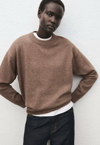 Maglione marrone lavorato a maglia con colletto rotondo, polsini e orlo a coste. Indossato sopra una t-shirt bianca, abbinato a jeans di denim scuro.